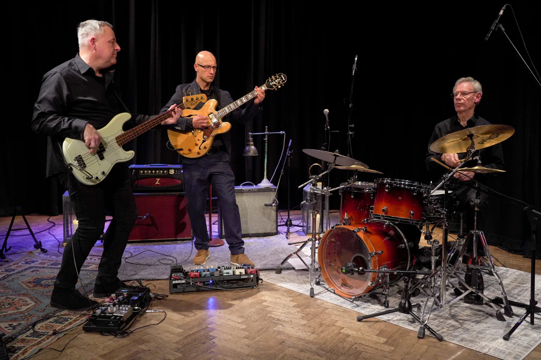Pete Roth Trio performance photo with guitar and drums in dramatic stage light.