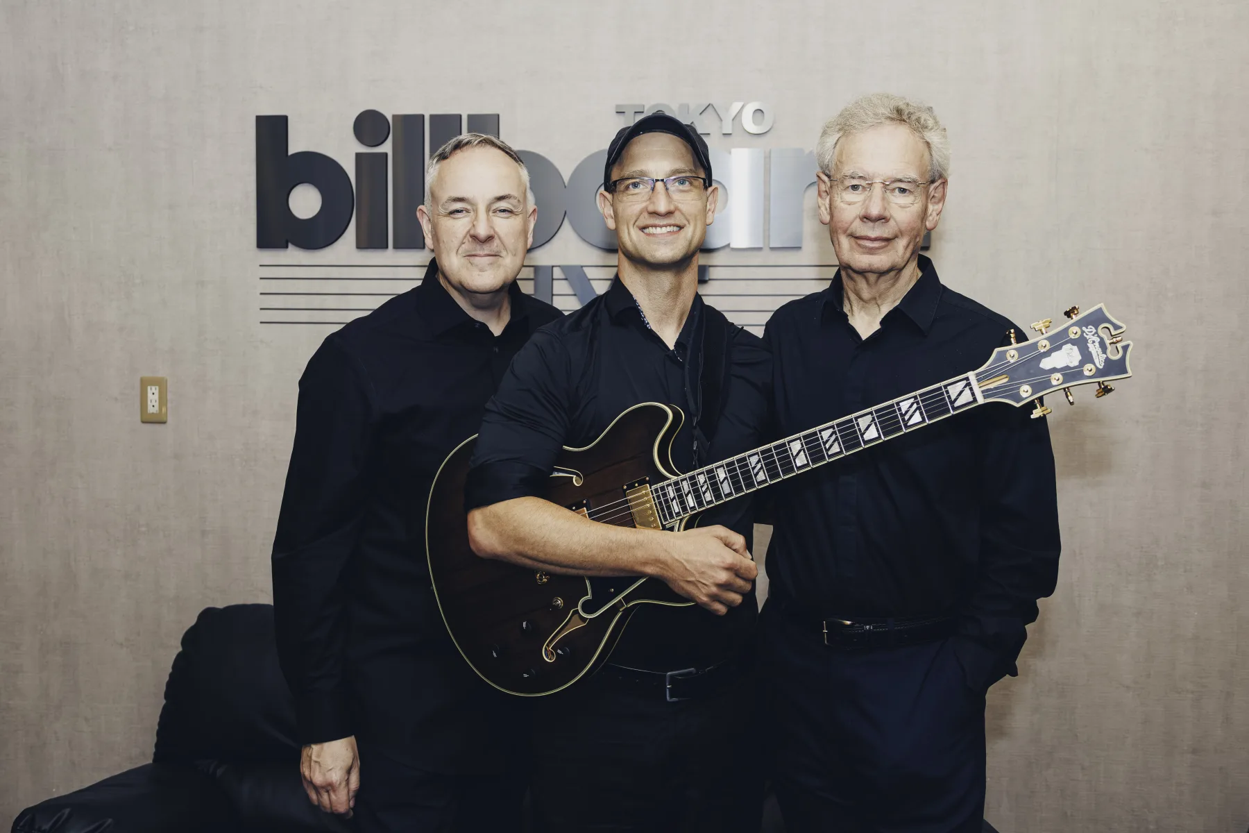 Pete Roth Trio standing together with guitar in front of the Billboard Live Tokyo wall.